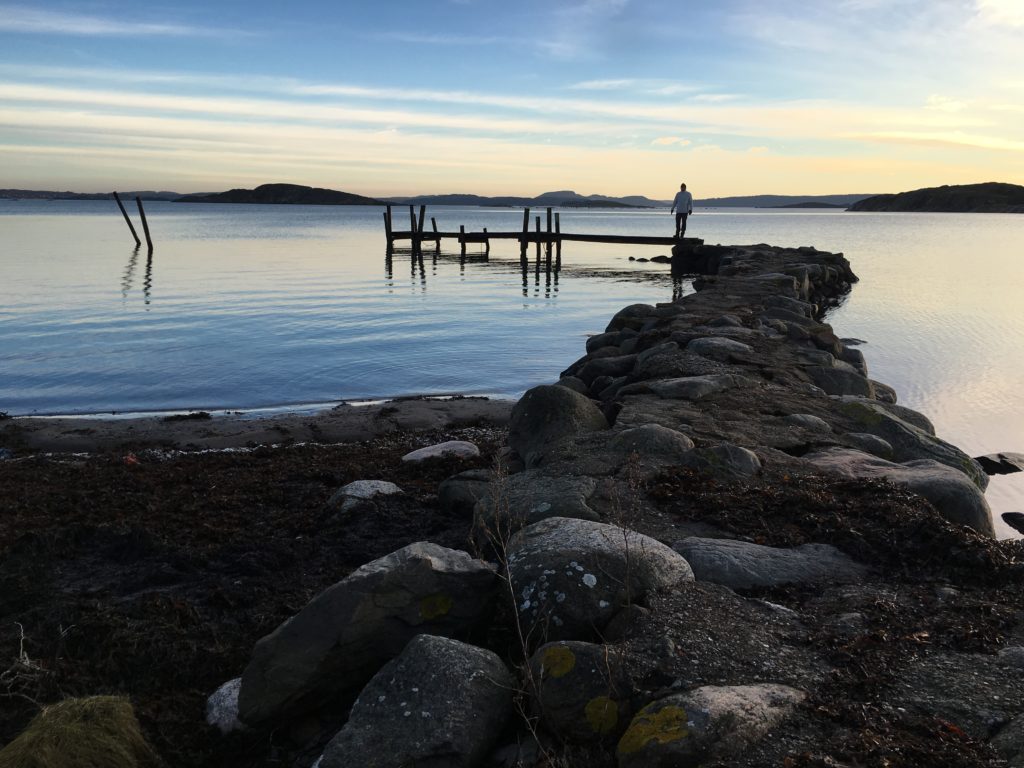 im Vordergrund befinden sich große Steine, die zu einem Weg verbunden sind. Im Hintergrund grenzt ein Holzsteg an die Steine und führt hinaus auf das ruhige Meer. Im Hintergrund sind kleine Berge und der blaue Himme mit den letzten Sonnenstrahen des Tages und leichten weissen Streifen an Wolken zu sehen.